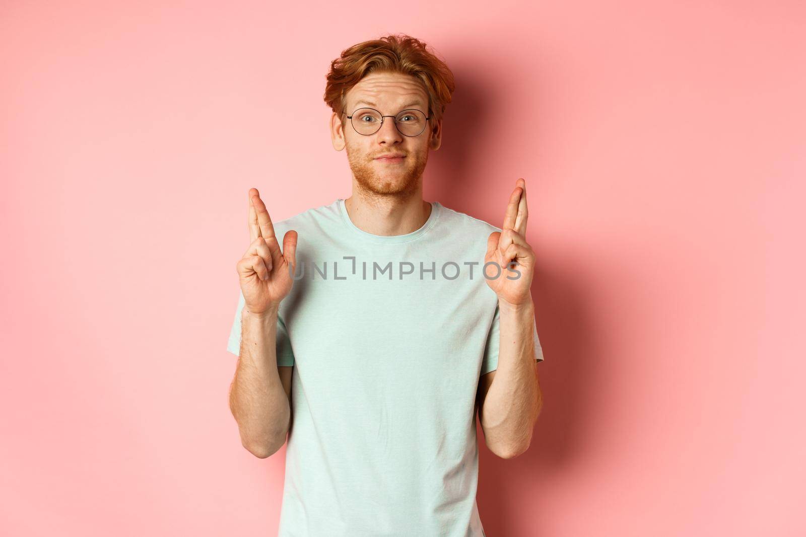 Attractive guy having faith in dreams, smiling hopeful and making wish with fingers crossed, feeling lucky, standing over pink background.
