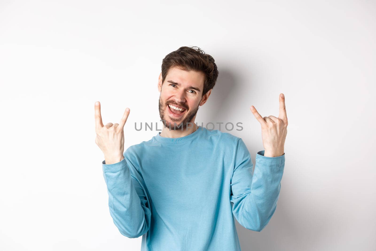 Excited handsome man celebrating, having fun and showing rock on horns gesture, enjoing party, smiling at camera, standing on white background by Benzoix