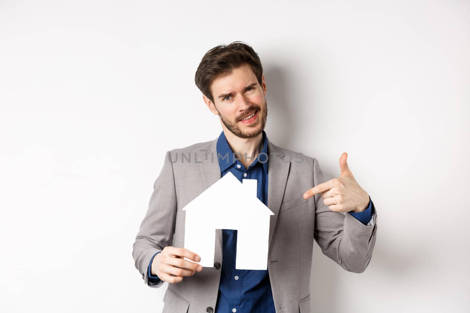 Real estate and insurance concept. Salesman in grey suit showing paper house cutout, selling property, smiling at camera, white background.