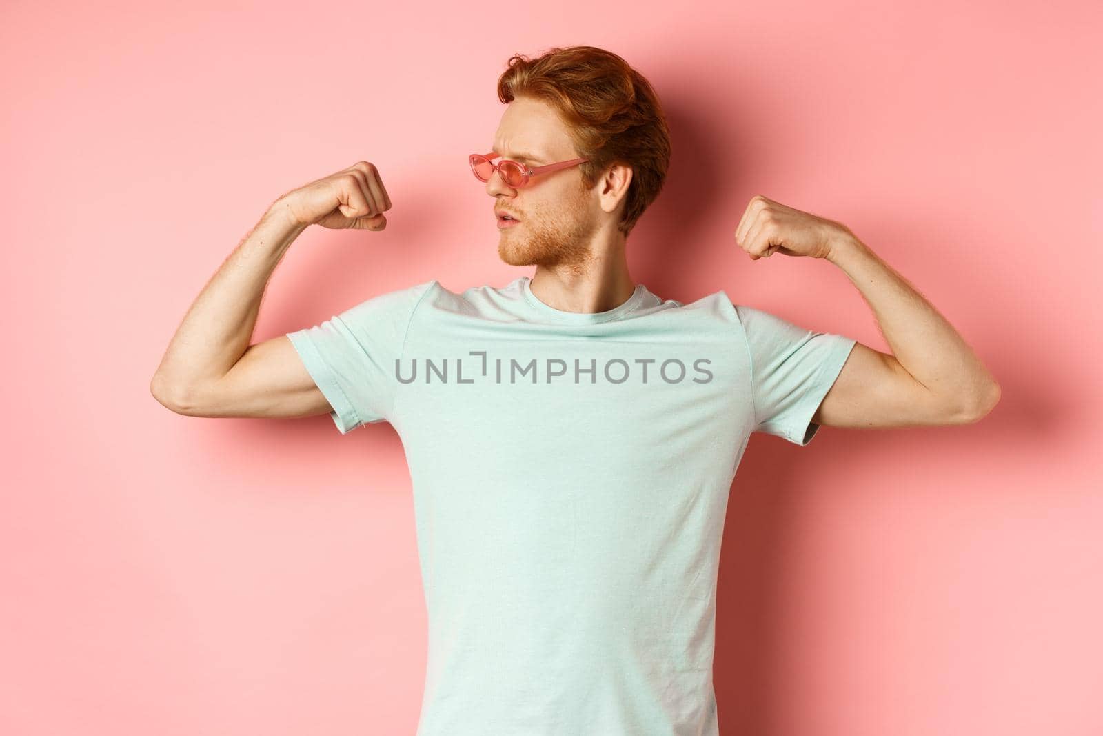 Confident young man with red hair, wearing summer sunglasses and t-shirt, showing strong and fit body muscles, flex biceps and staring cool at camera, pink background by Benzoix