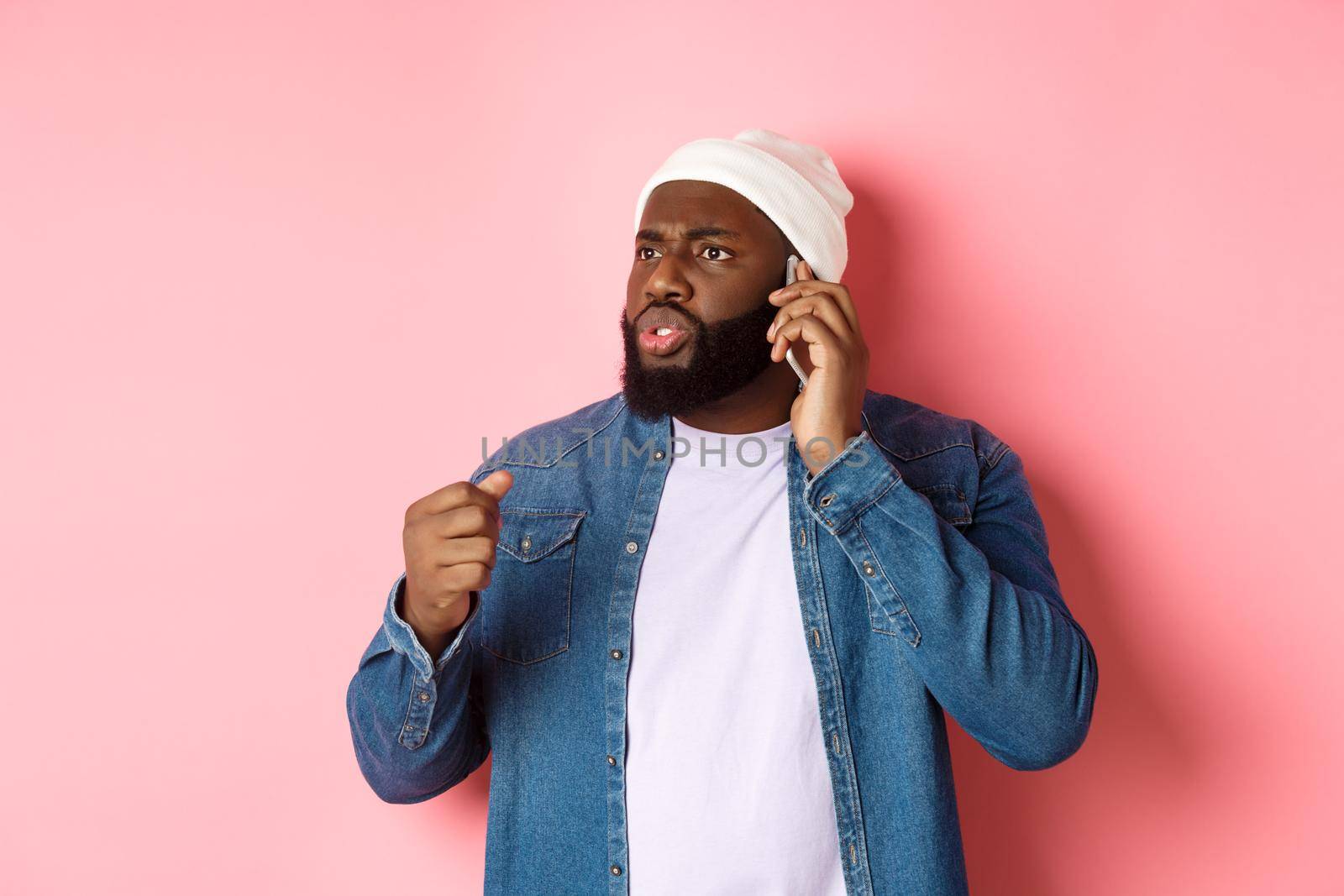 Angry african-american man talking on phone and threaten someone with raised fist, looking bothered at person, standing over pink background.
