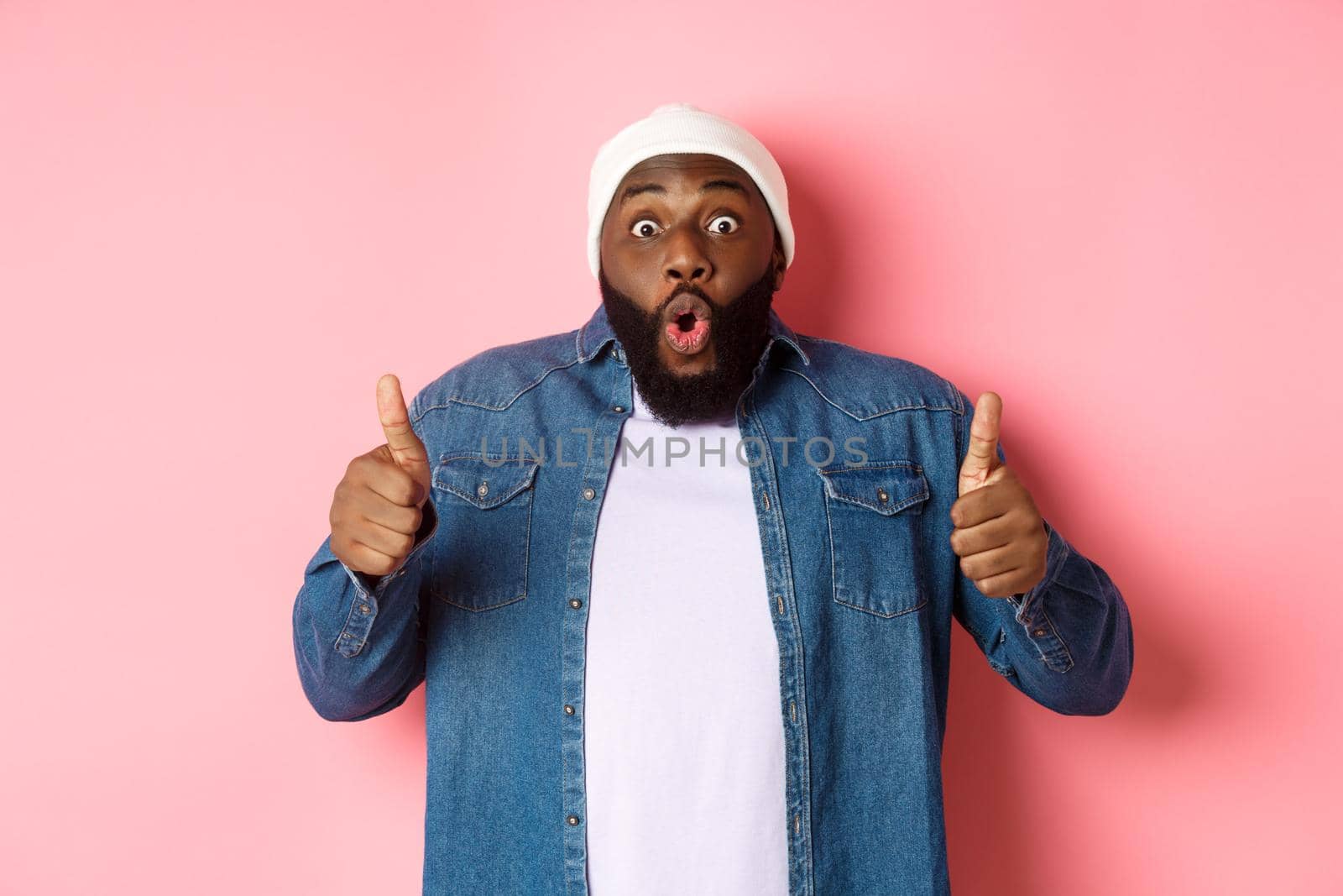 Impressed african-american hipster guy showing thumbs-up, approve and like offer, praising great choice, standing over pink background by Benzoix