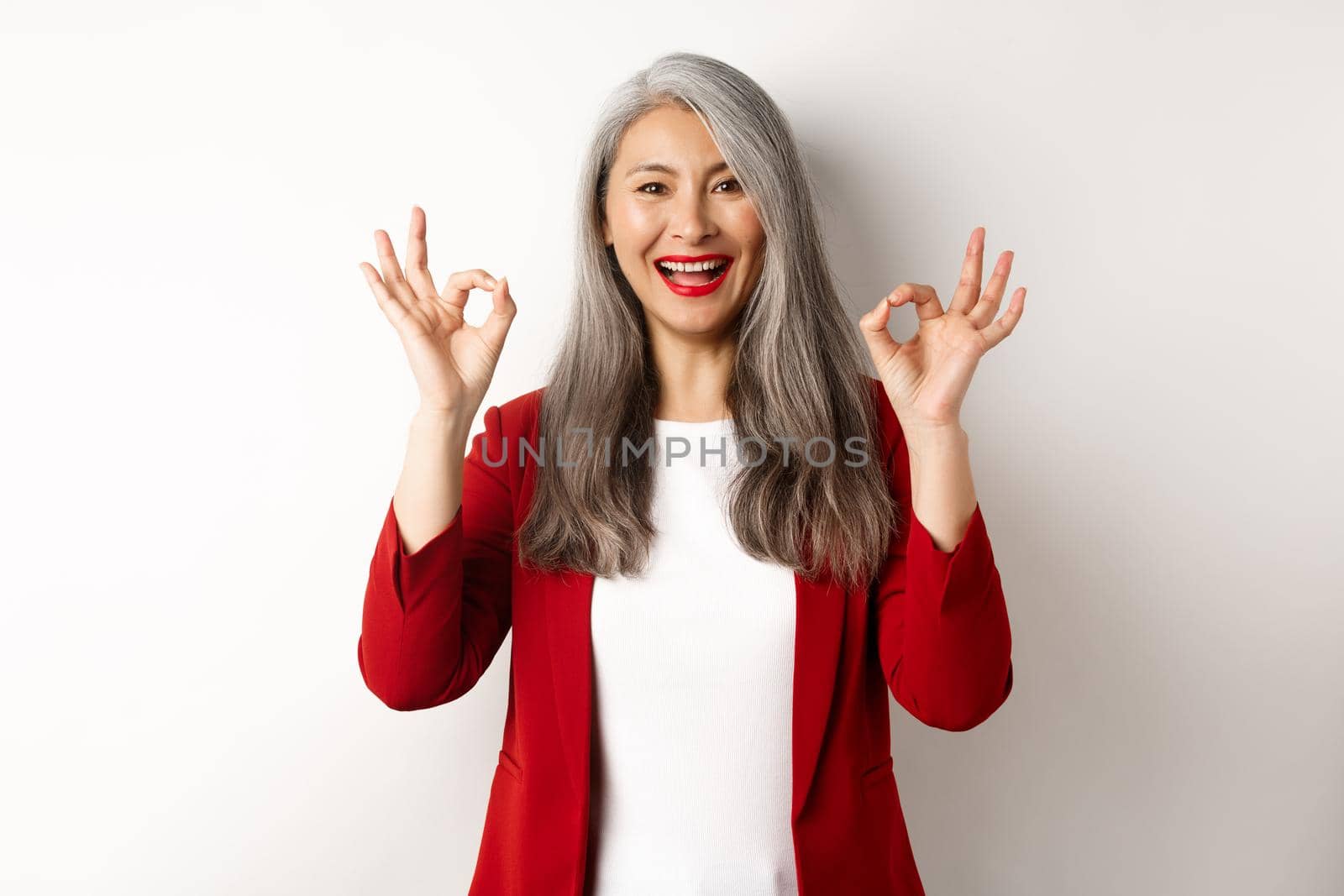 Beautiful senior asian woman in red blazer, smiling and showing okay signs, approve and say yes, standing over white background.