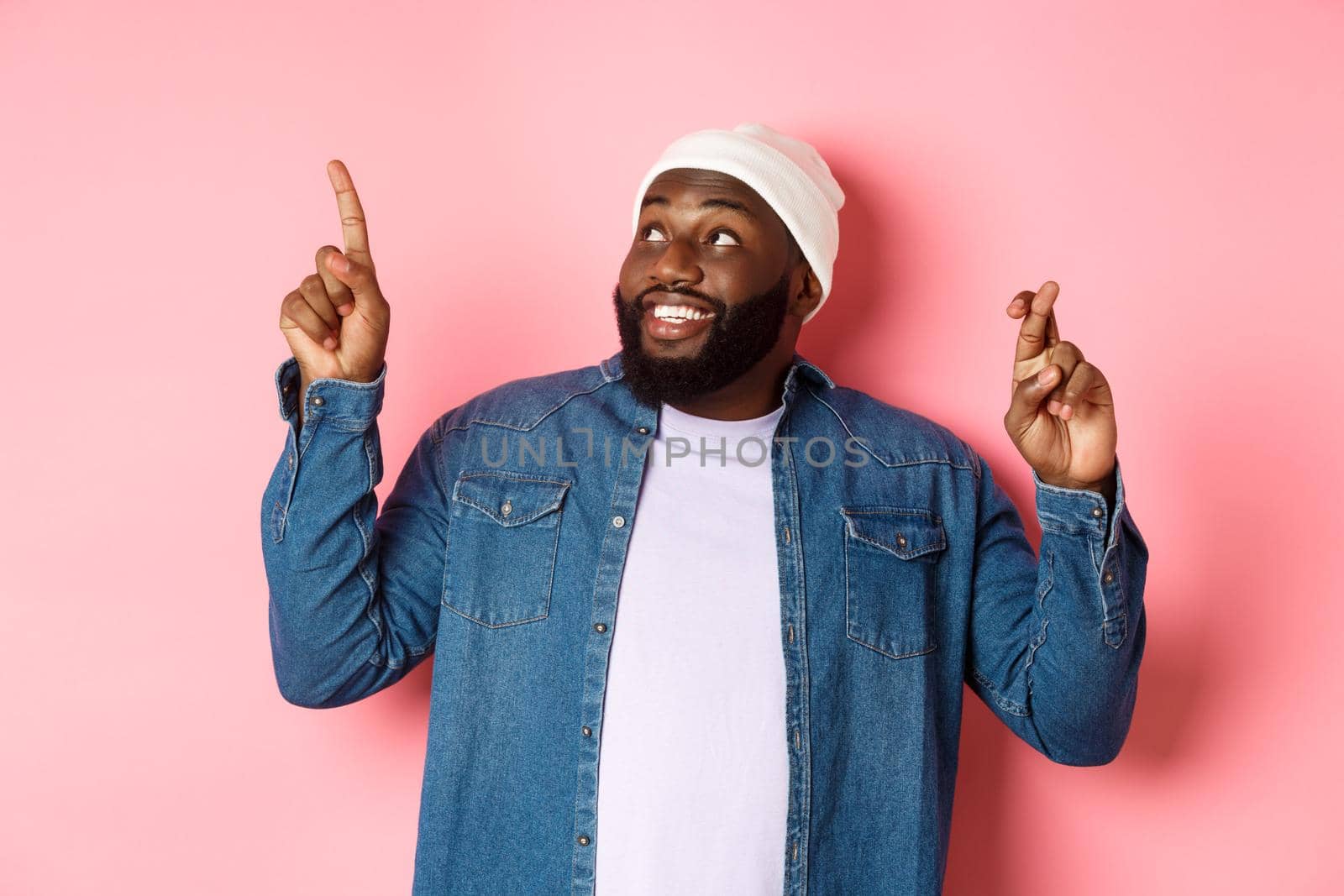 Hopeful african-american man making a wish, cross fingers for good luck, pointing and looking at upper left corner, dreaming about buying something, pink background.