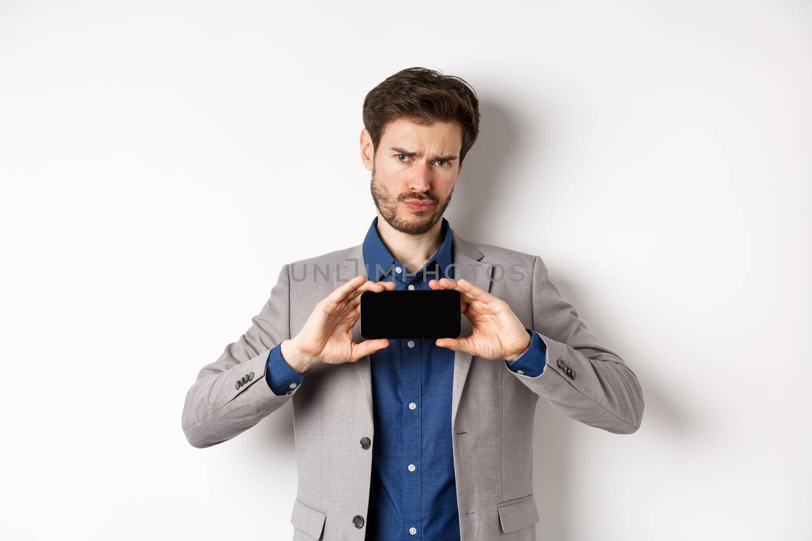 E-commerce and online shopping concept. Hesitant frowning man showing empty smartphone screen, standing on white background.