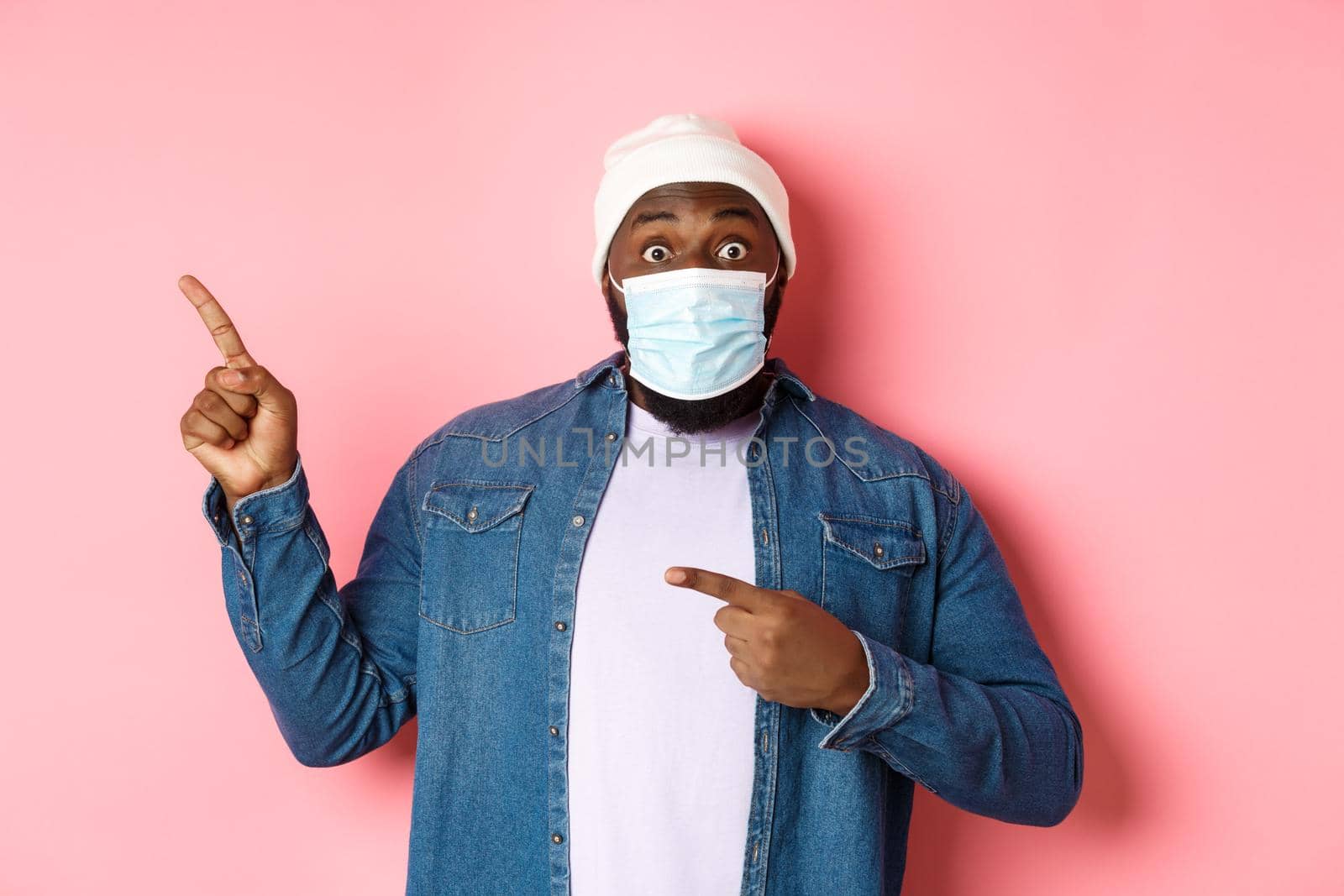 Coronavirus, lifestyle and global pandemic concept. Amazed black man in face mask showing advertisement, pointing at upper left corner and look excited, pink background.