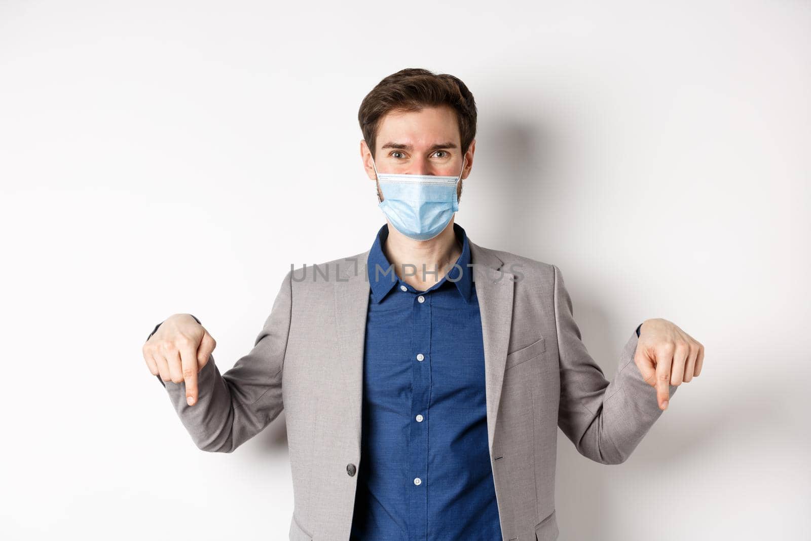 Covid-19, pandemic and business concept. Handsome businessman in medical mask and suit, pointing fingers down and showing company logo, white background by Benzoix