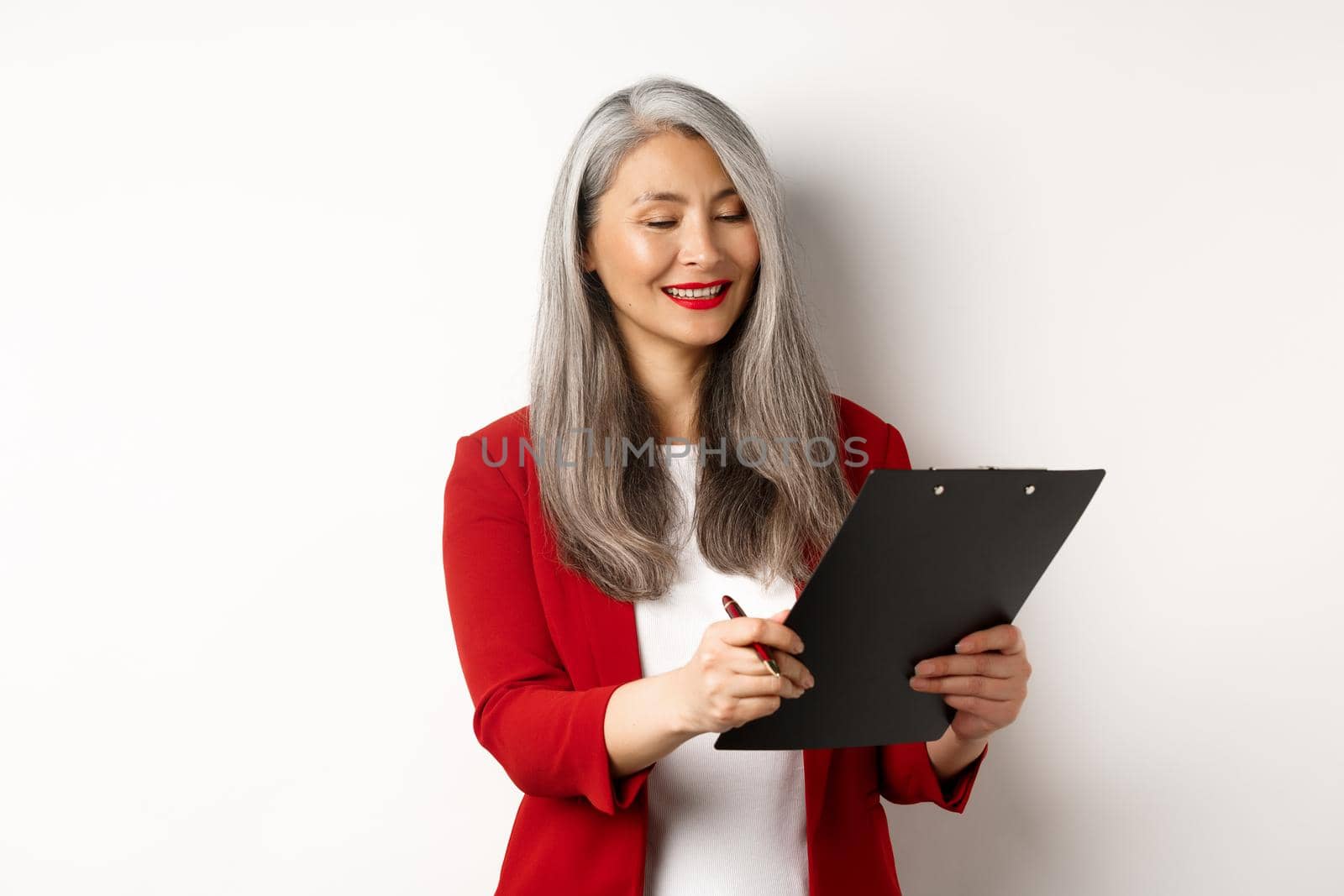 Successful asian businesswoman signing a deal, looking satisfied at documents on clipboard and smiling, standing over white background.