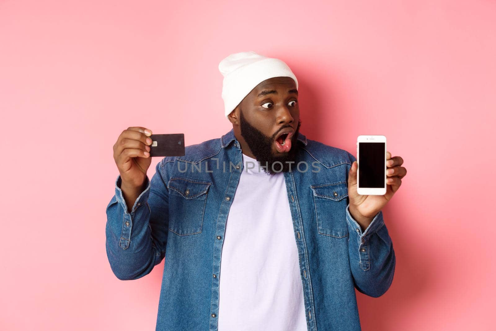 Online shopping. Impressed and surprised Black man showing credit card, staring at smartphone screen, demonstrate app, standing over pink background.