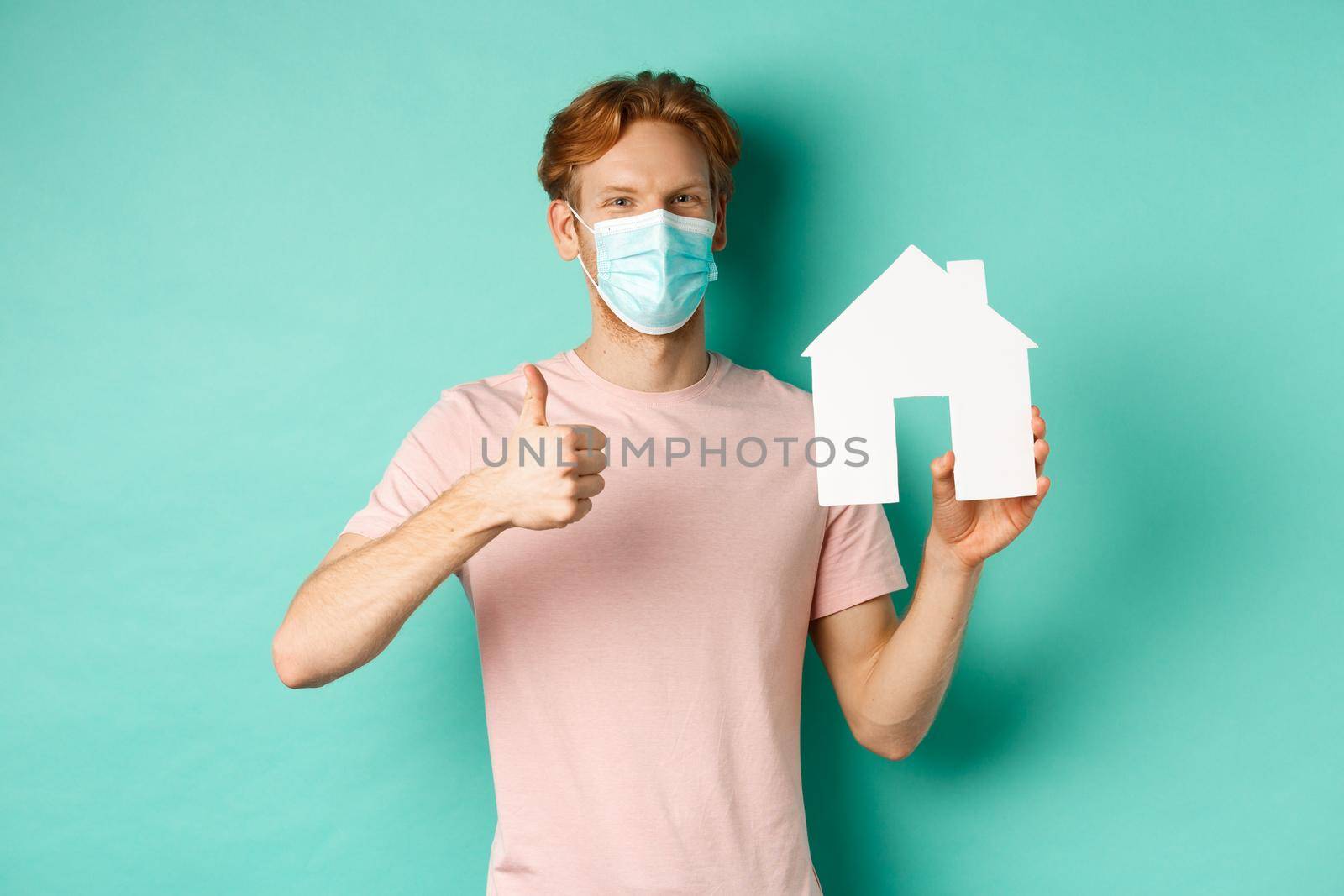 Covid-19 and real estate concept. Cheerful guy in face mask showing house cutout and thumbs-up, standing over turquoise background.