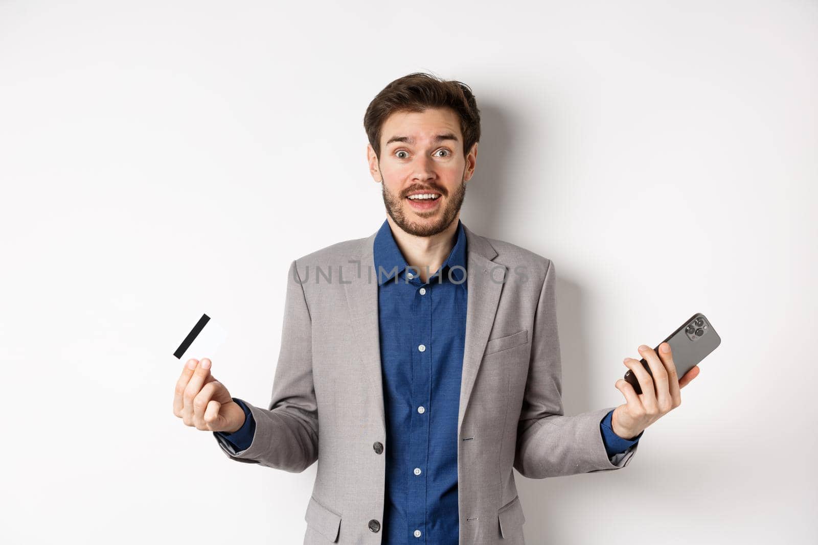 Online shopping. Surprised businessman holding plastic credit card and smartphone, making money in internet, standing in suit against white background.