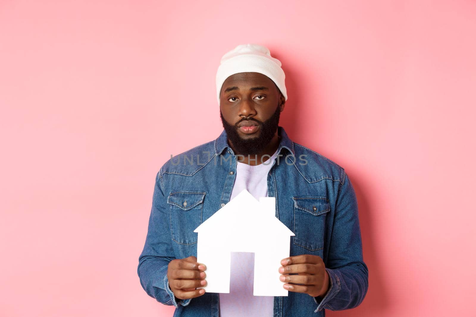Real estate concept. Sad and tired Black man staring unamused at camera, holding paper house model, standing over pink background by Benzoix