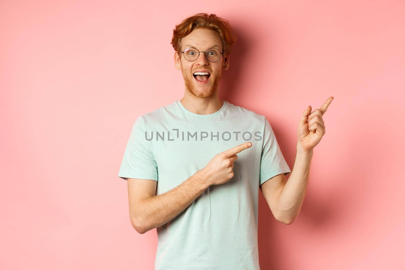 Amazed redhead man in glasses and t-shirt showing special promotion, pointing fingers at upper right corner and staring excited at camera, standing over pink background by Benzoix