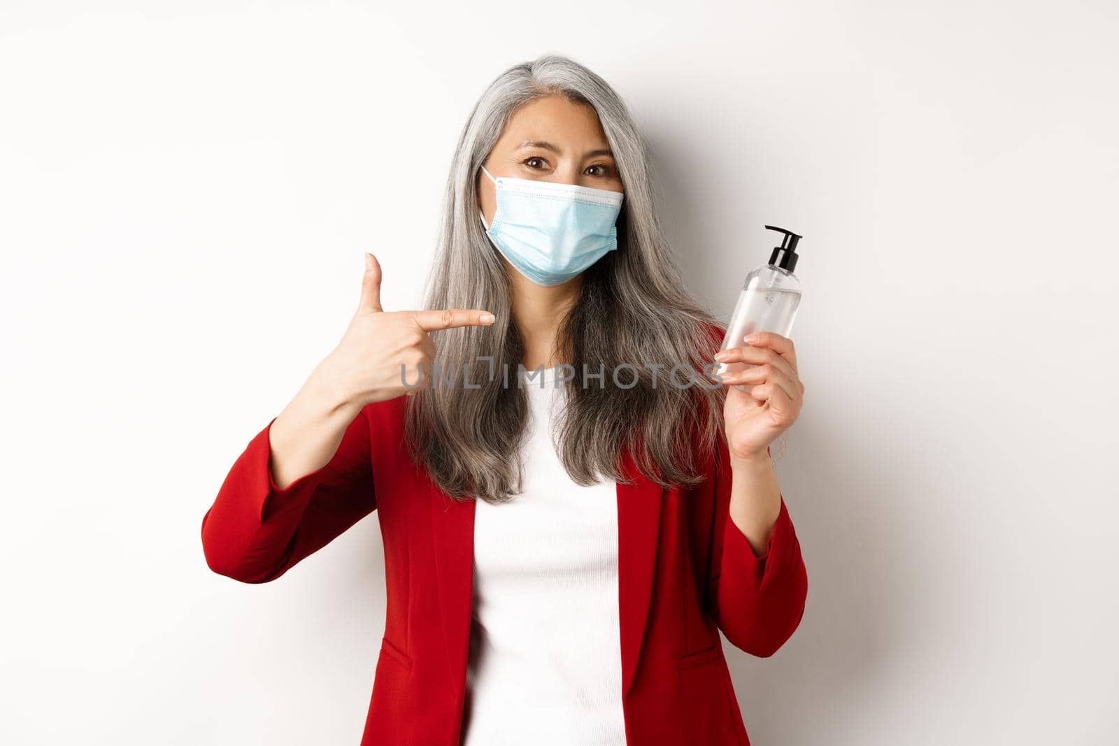 Covid, pandemic and business concept. Asian female manager in face mask, pointing finger at hand sanitizer, recommending antiseptic, white background by Benzoix