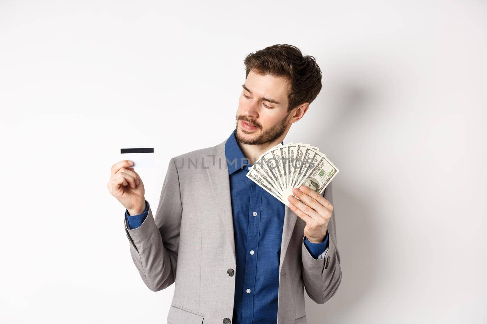 Successful man holding dollar bills and looking at plastic credit card, pleased with bank, standing on white background in business suit.