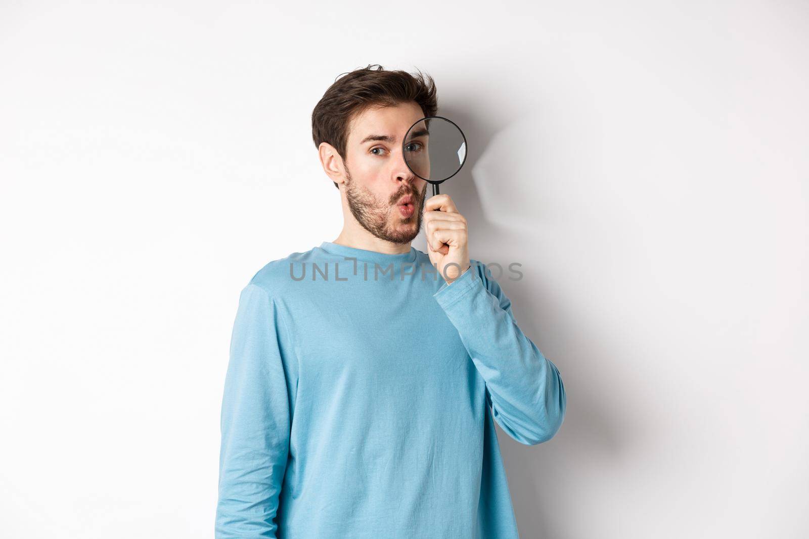 Surprised man found something, looking through magnifying glass with amazed face, standing over white background by Benzoix