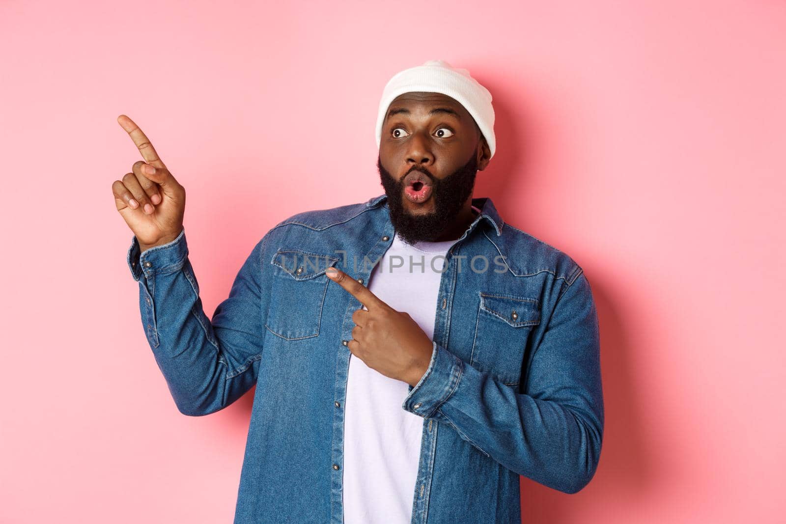 Impressed hipster black man in beanie and denim shirt, looking and pointing at upper left corner logo, staring amazed, standing over pink background.