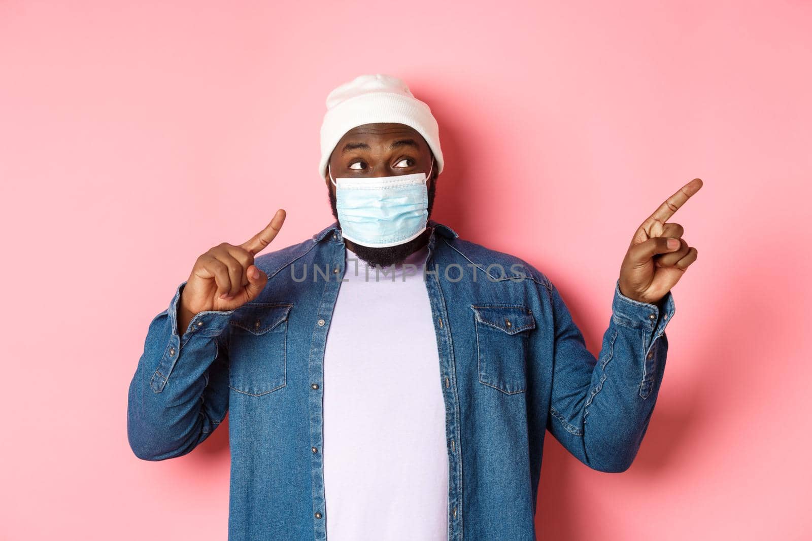 Covid-19, lifestyle and lockdown concept. Amused Black man in medical mask, looking and pointing right at copy space, standing over pink background by Benzoix