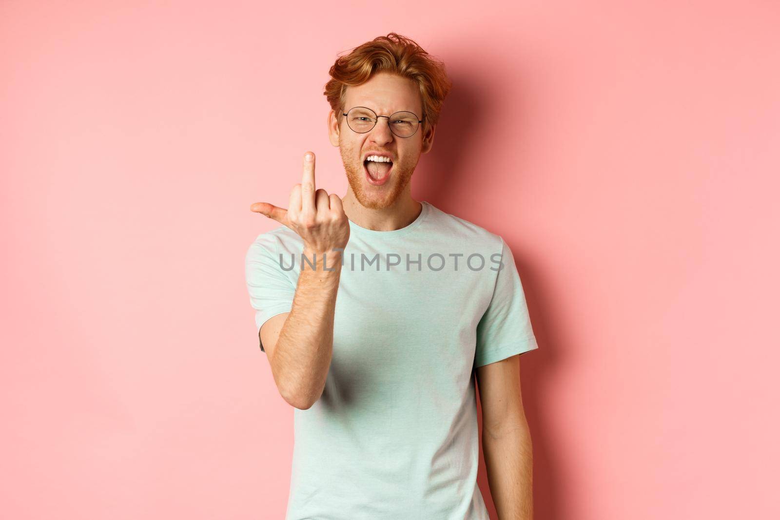 Arrogant and rude redhead man in glasses dont give a fuck, showing middle fingers at camera and frowning, standing over pink background.