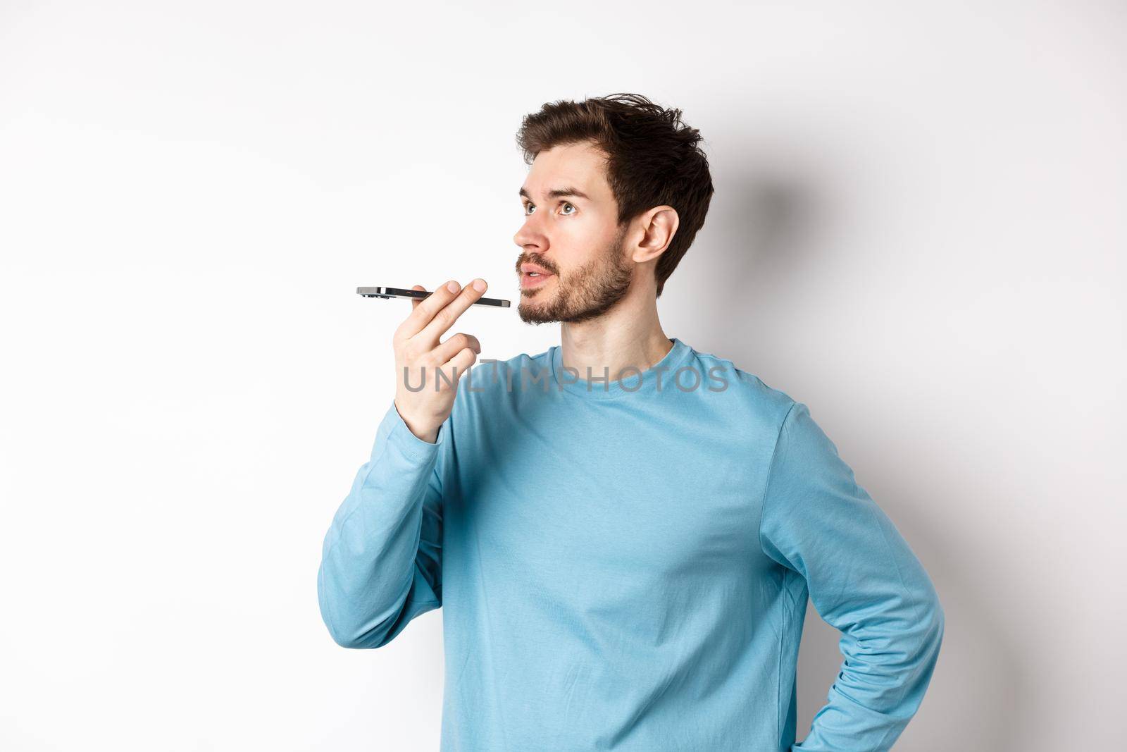 Handsome young man recording voice message on smartphone, holding phone near lips and talking. Guy using translator app on mobile phone, white background by Benzoix