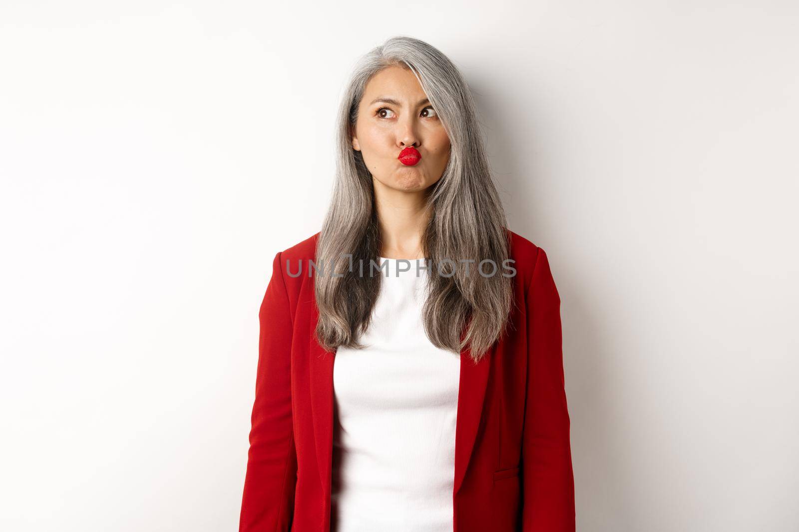 Indecisive asian businesswoman pucker lips and looking thoughtful at upper left corner, standing over white background.