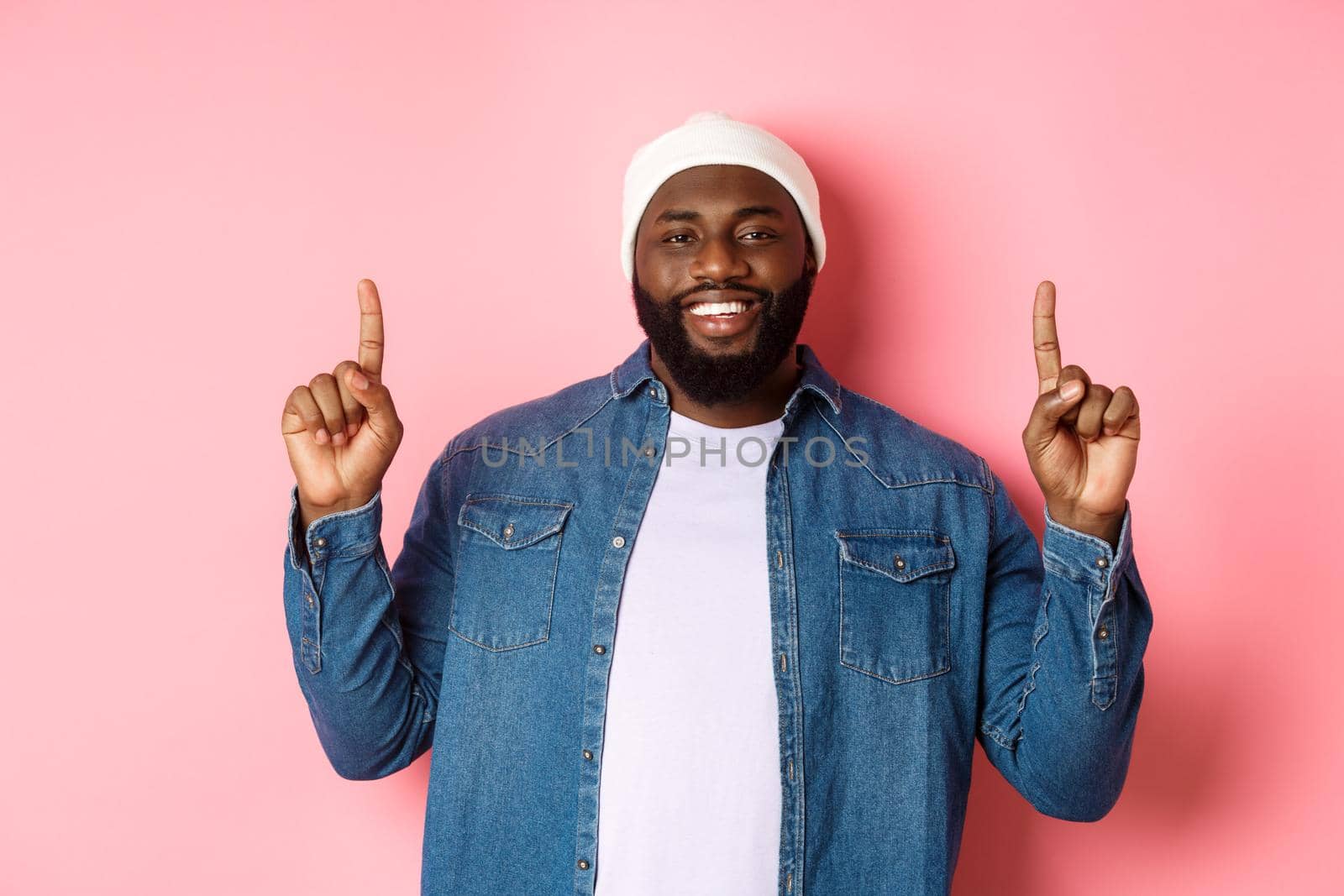 Handsome happy african-american man pointing fingers up, smiling and showing promo offer, wearing hipster beanie and denim shirt, pink background.