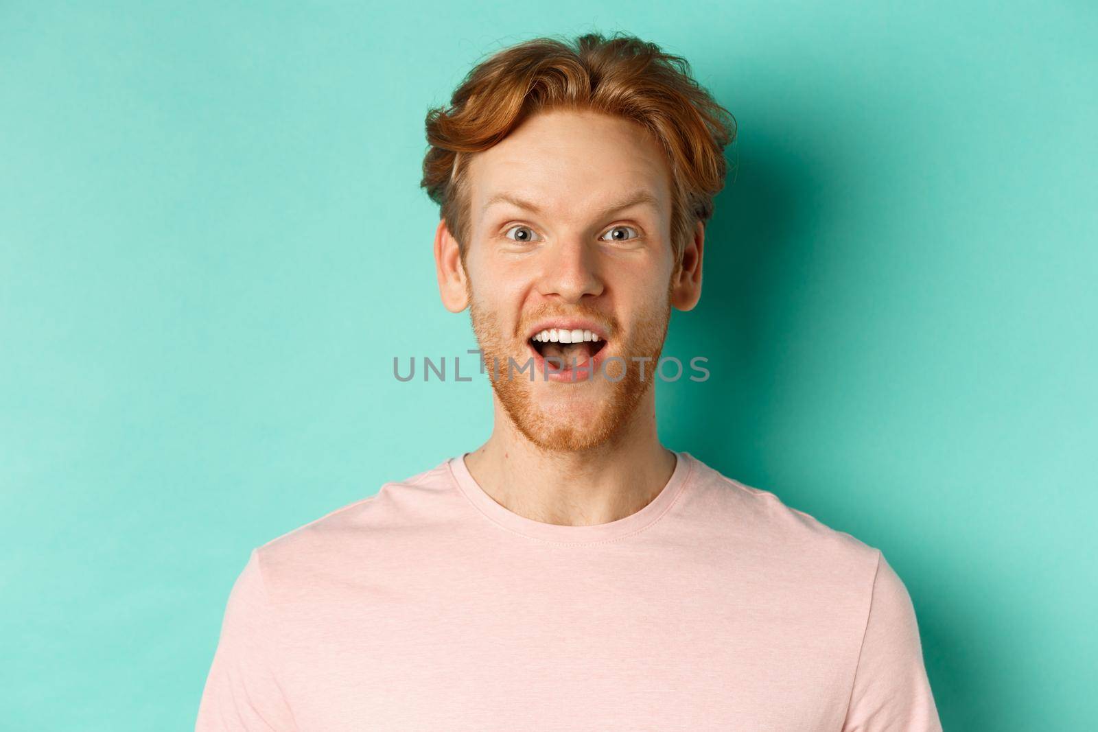 Close up of redhead bearded man raising eyebrows and looking surprised at camera, checking out promotion, standing over mint background.