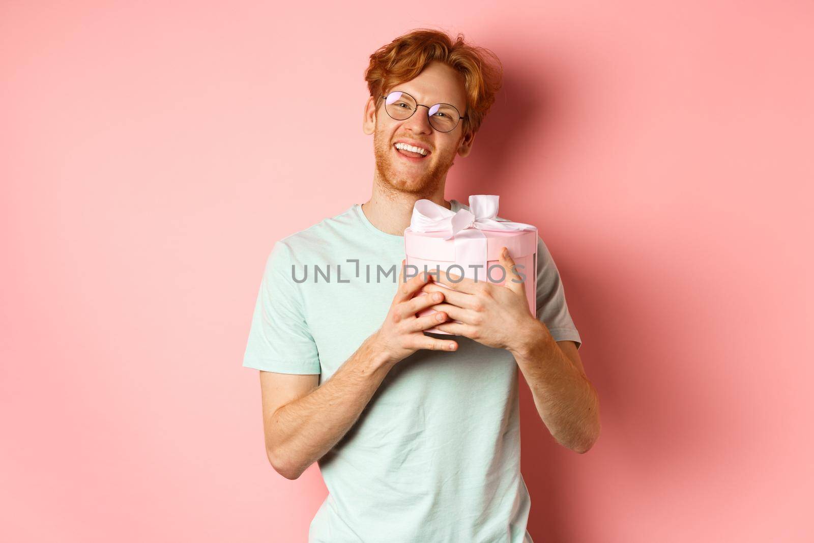 Valentines day and romance concept. Happy redhead boyfriend receive romantic gift, hugging box with present and saying thank you, smiling grateful, pink background by Benzoix