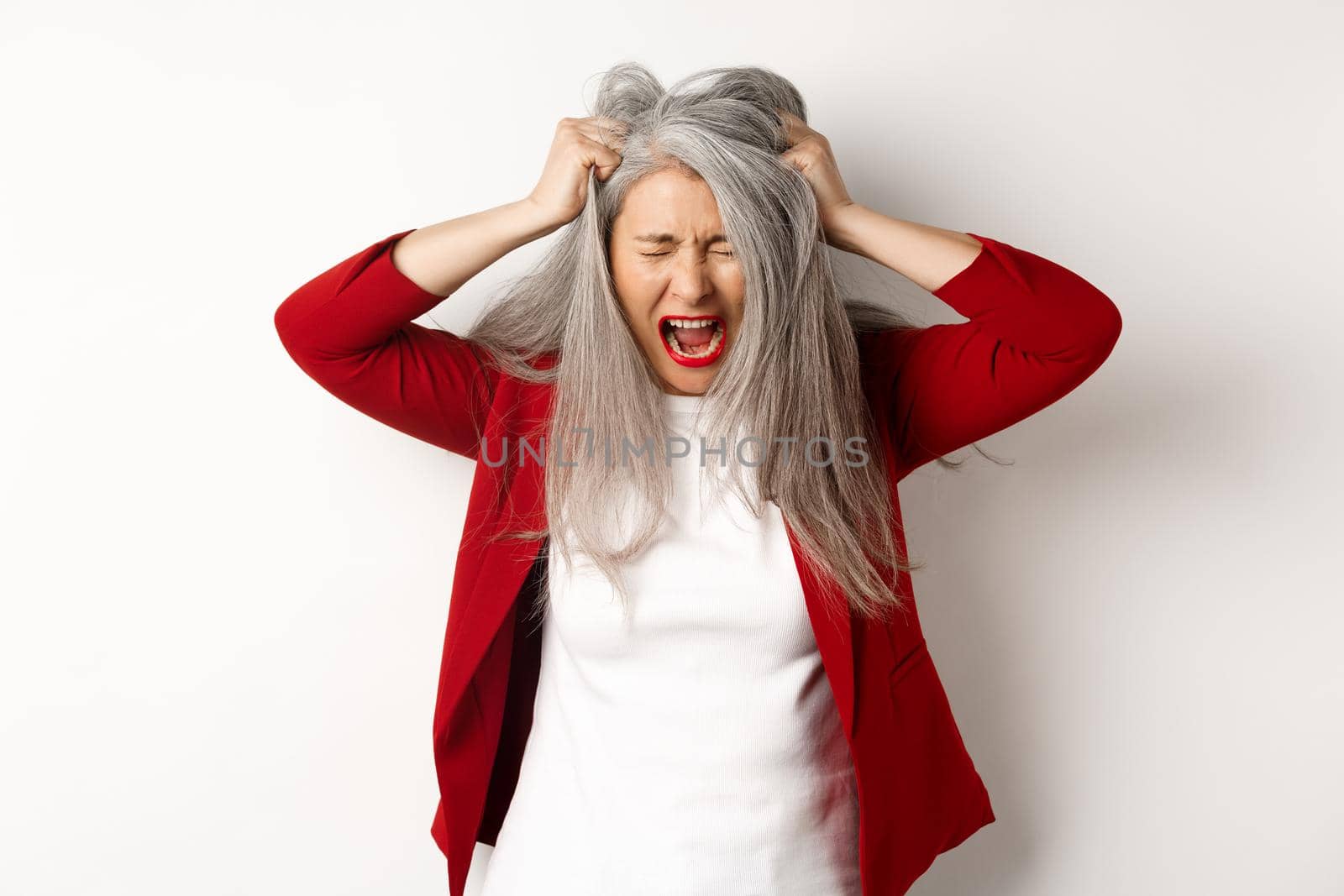 Distressed and pissed-off asian senior woman pulling hair and screaming, standing upset over white background. Copy space