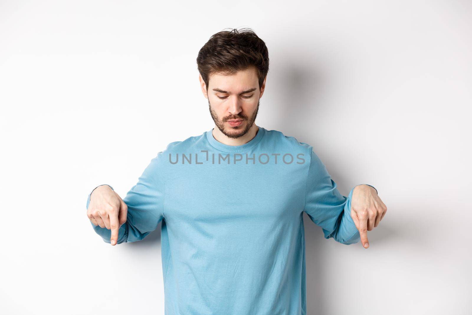 Handsome caucasian man with beard, wearing blue shirt, looking and pointing down at interesting logo banner, standing on white background.