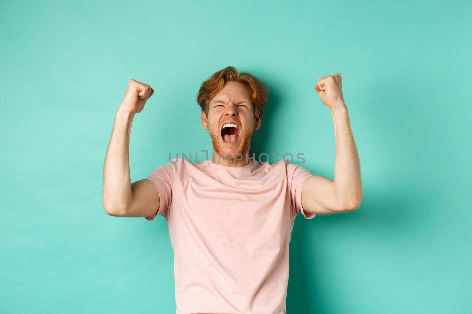 Young redhead man triumphing as watching sport game, making bet on winning team and rejoicing, screaming with joy and satisfaction, standing over turquoise background.
