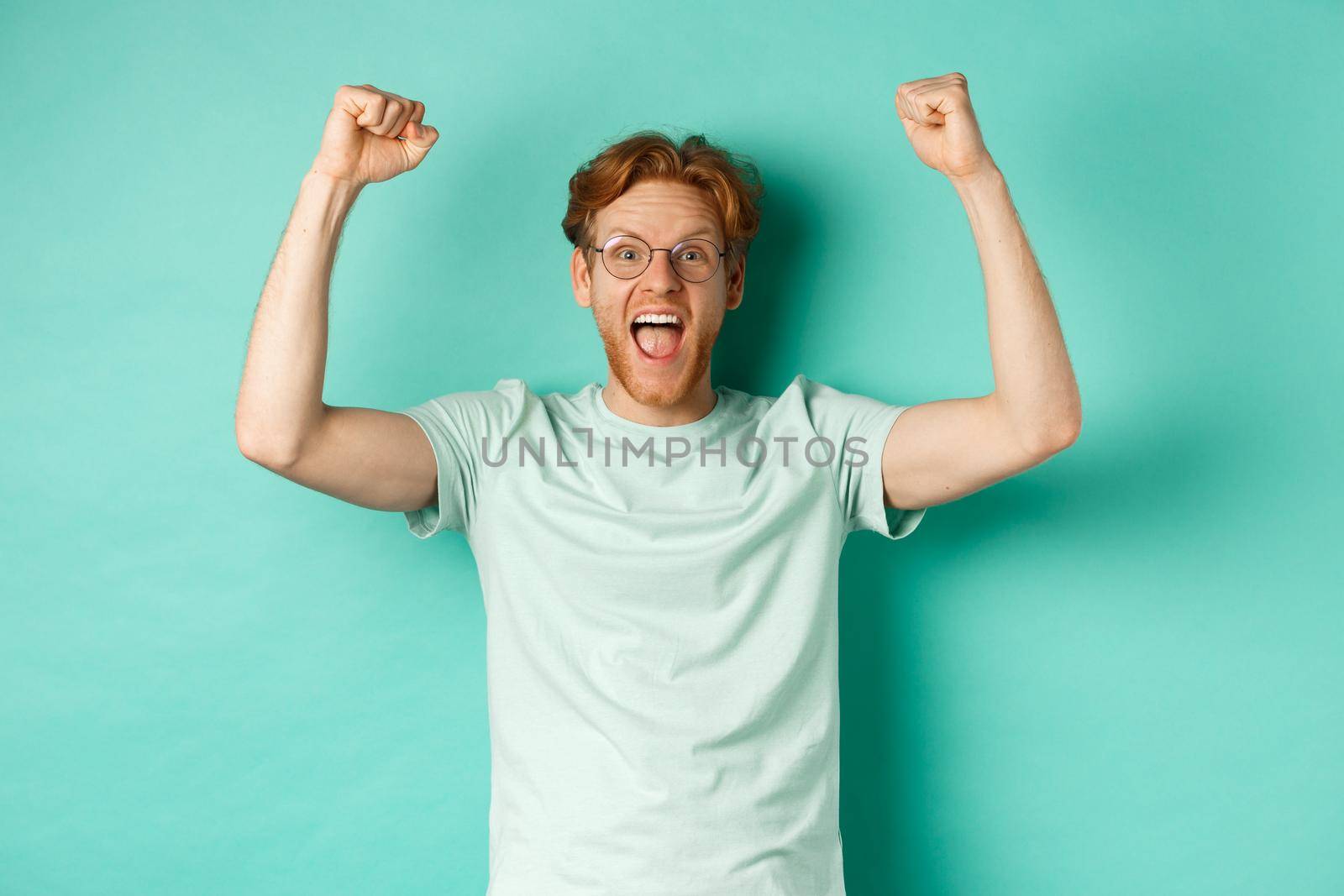 Young redhead man feeling like champion, raising hands up in fist pump gesture and shouting yes with joy, winning prize, triumphing of success, standing over mint background.