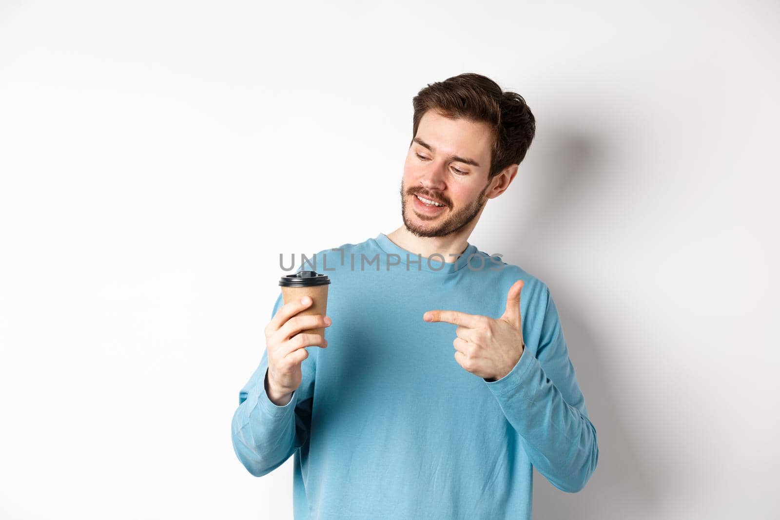 Handsome caucasian man pointing finger at coffee cup, recommending cafe, standing on white background satisfied.