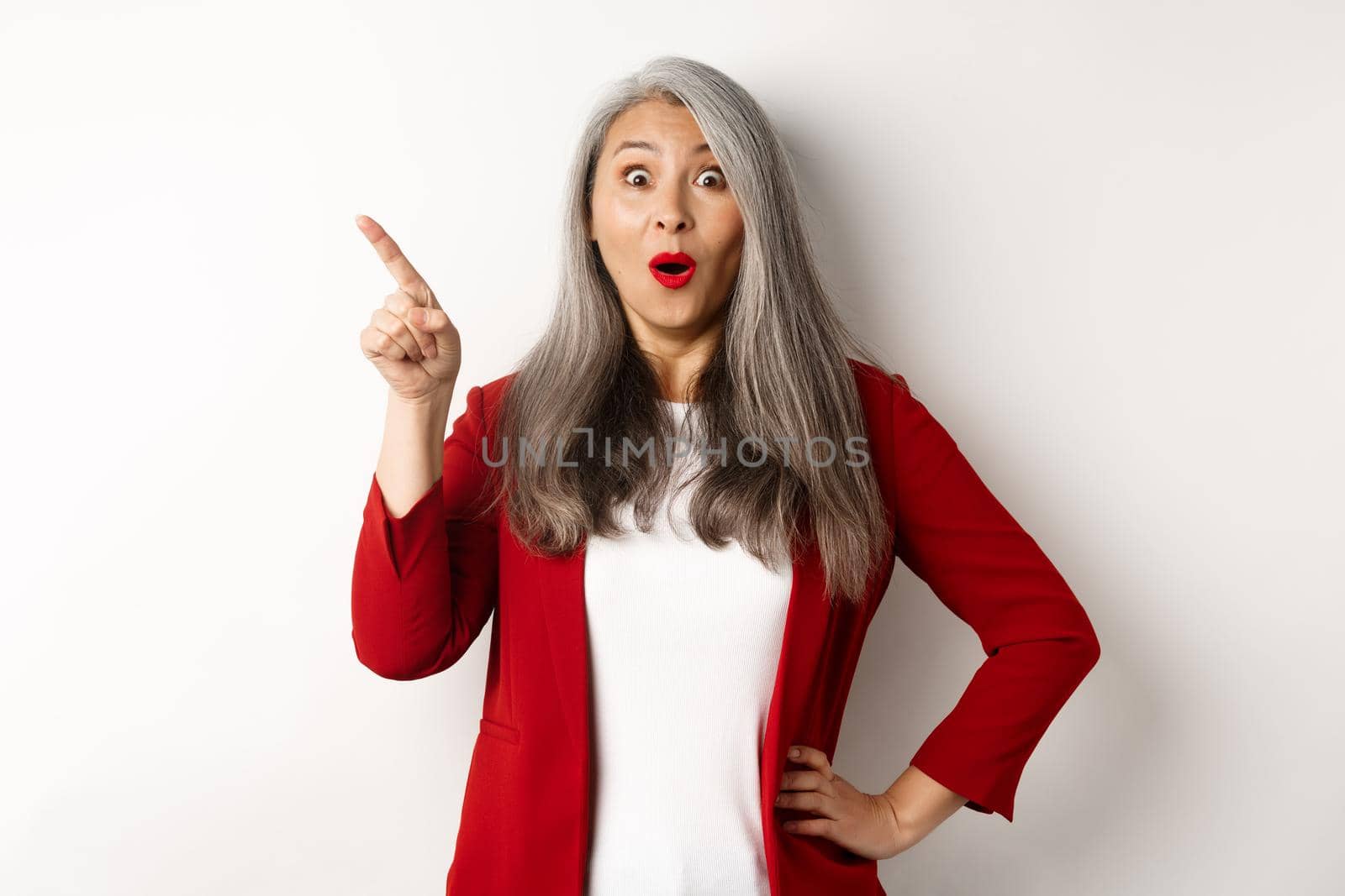 Impressed asian female entrepreneur in red blazer, pointing upper left corner and staring amazed at camera, checking out advertisement, white background by Benzoix