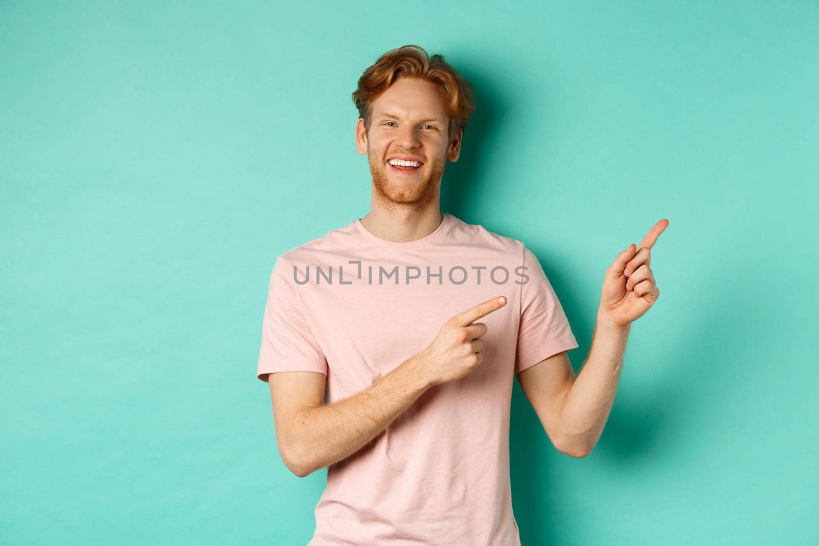 Handsome male model with red messy hair showing advertisement on copy space, pointing at upper right corner and smiling happy, standing over turquoise background.