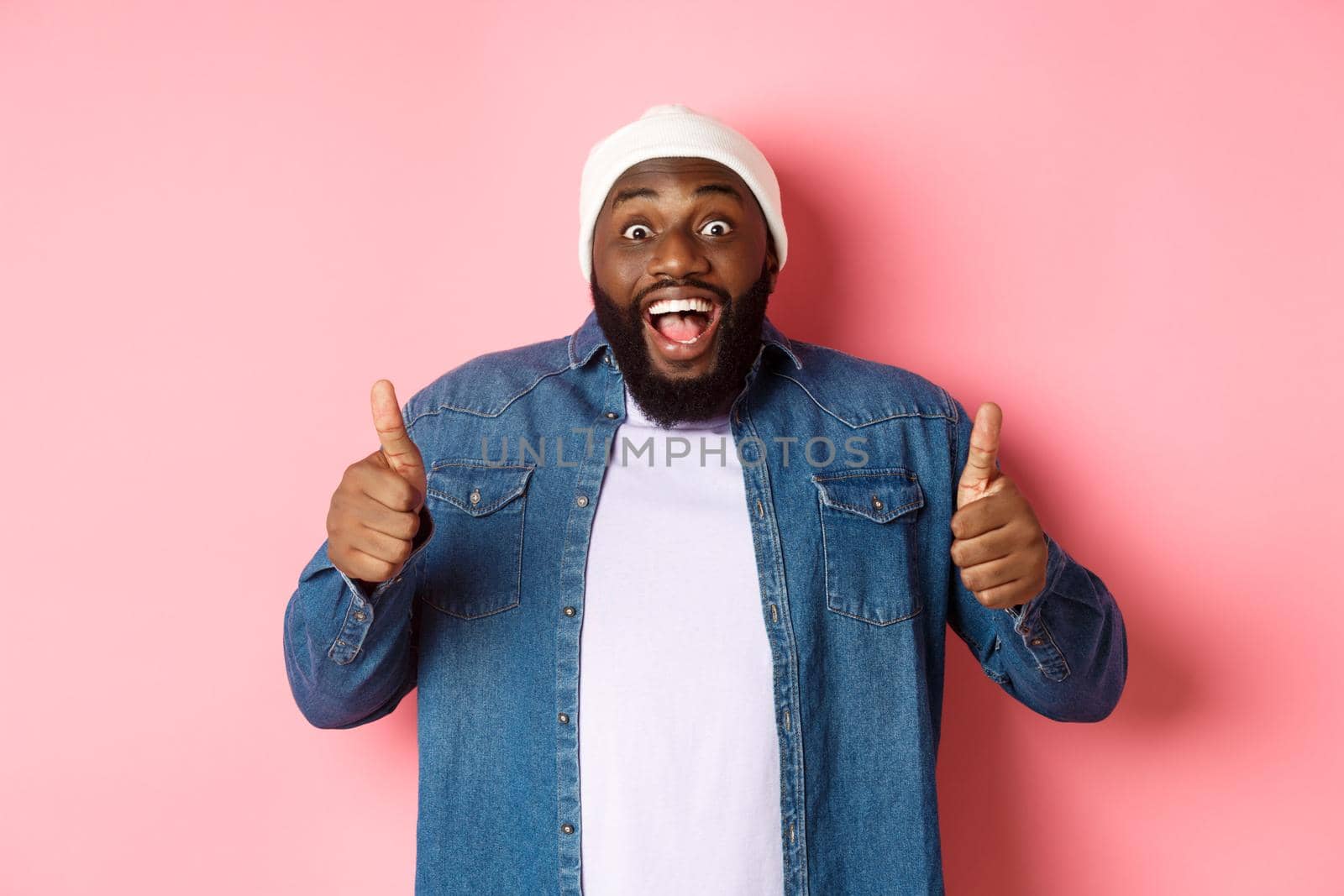 Impressed african-american hipster guy showing thumbs-up, approve and like offer, praising great choice, standing over pink background.