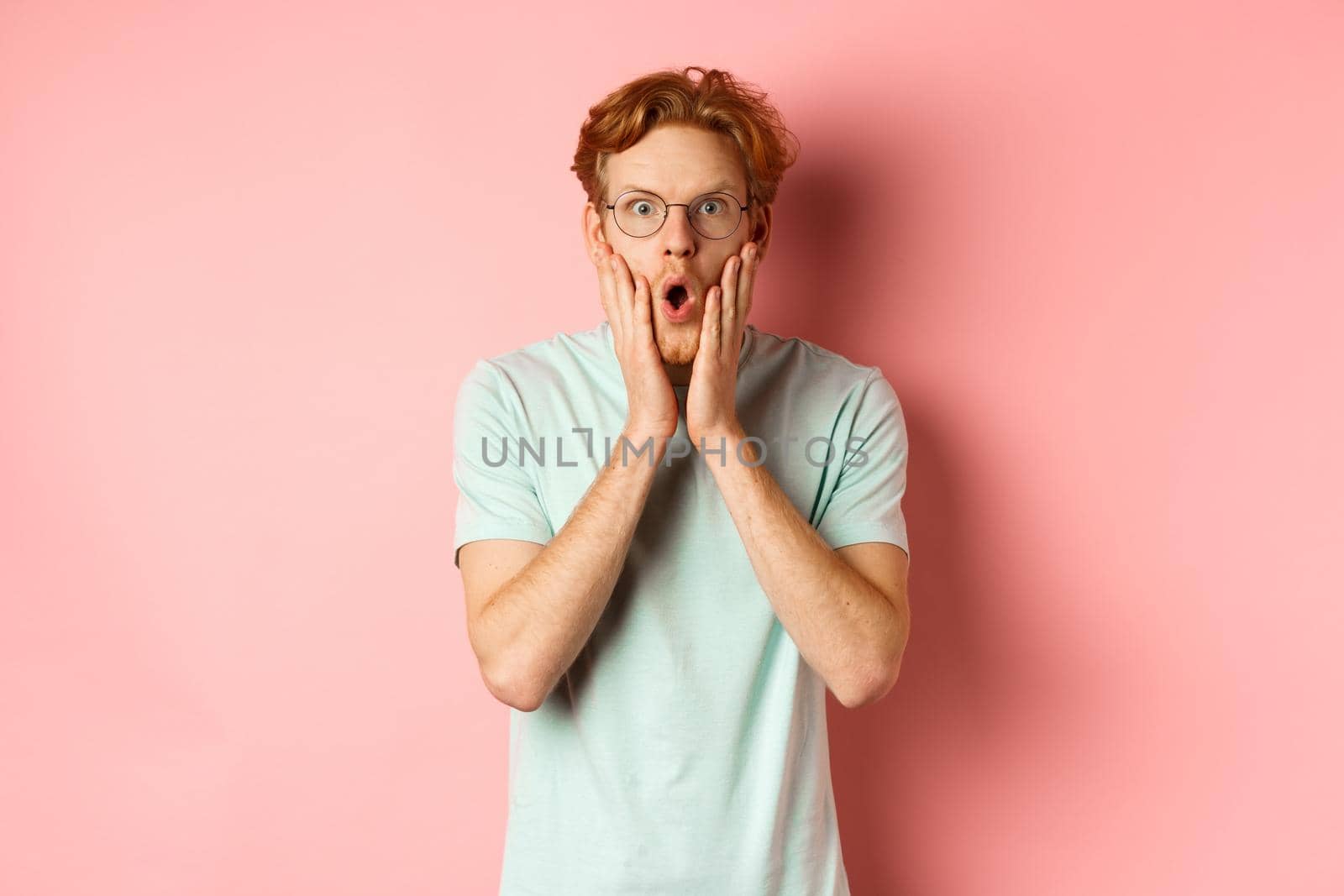 Shocked redhead man gossiping, staring impressed at camera and touching face, standing in glasses against pink background.