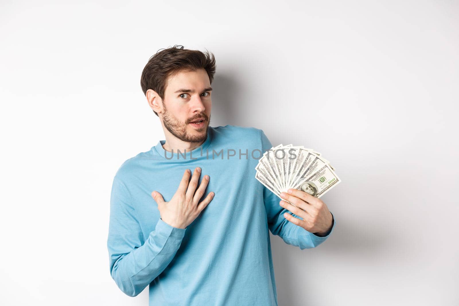 Surprised man holding money, looking amazed at camera, touching chest and gasping, standing in blue sweatshirt over white background.