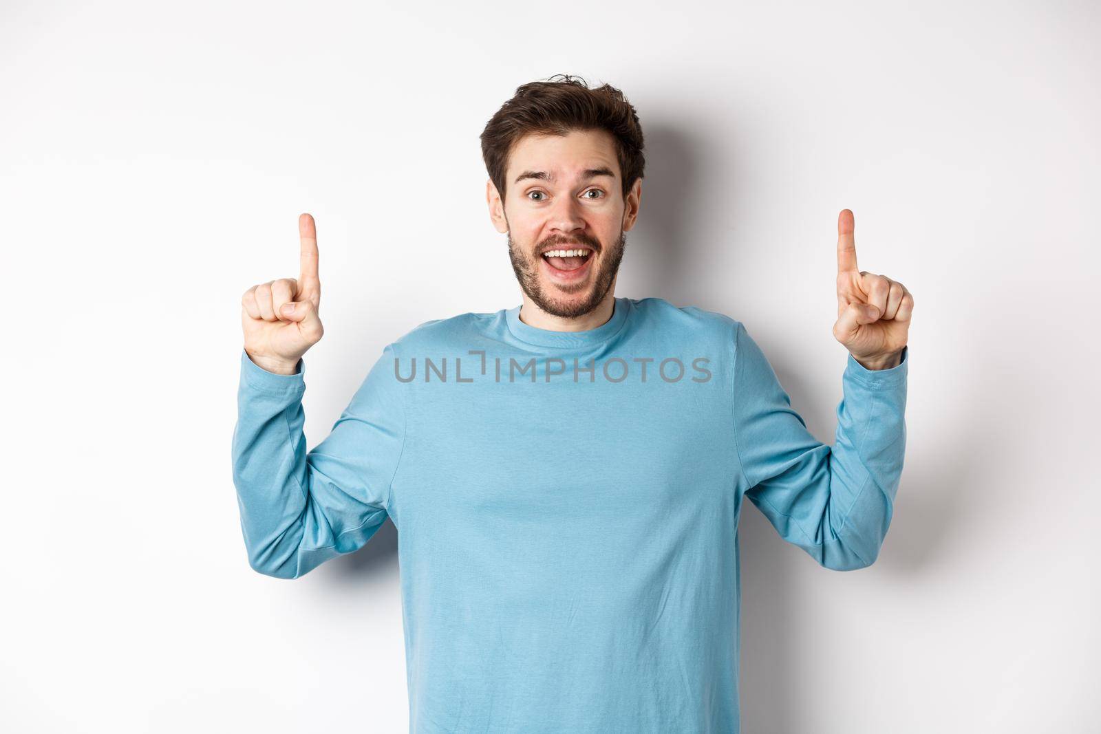 Cheerful young man showing advertisement with happy smile, pointing fingers up at awesome logo banner, standing on white background.