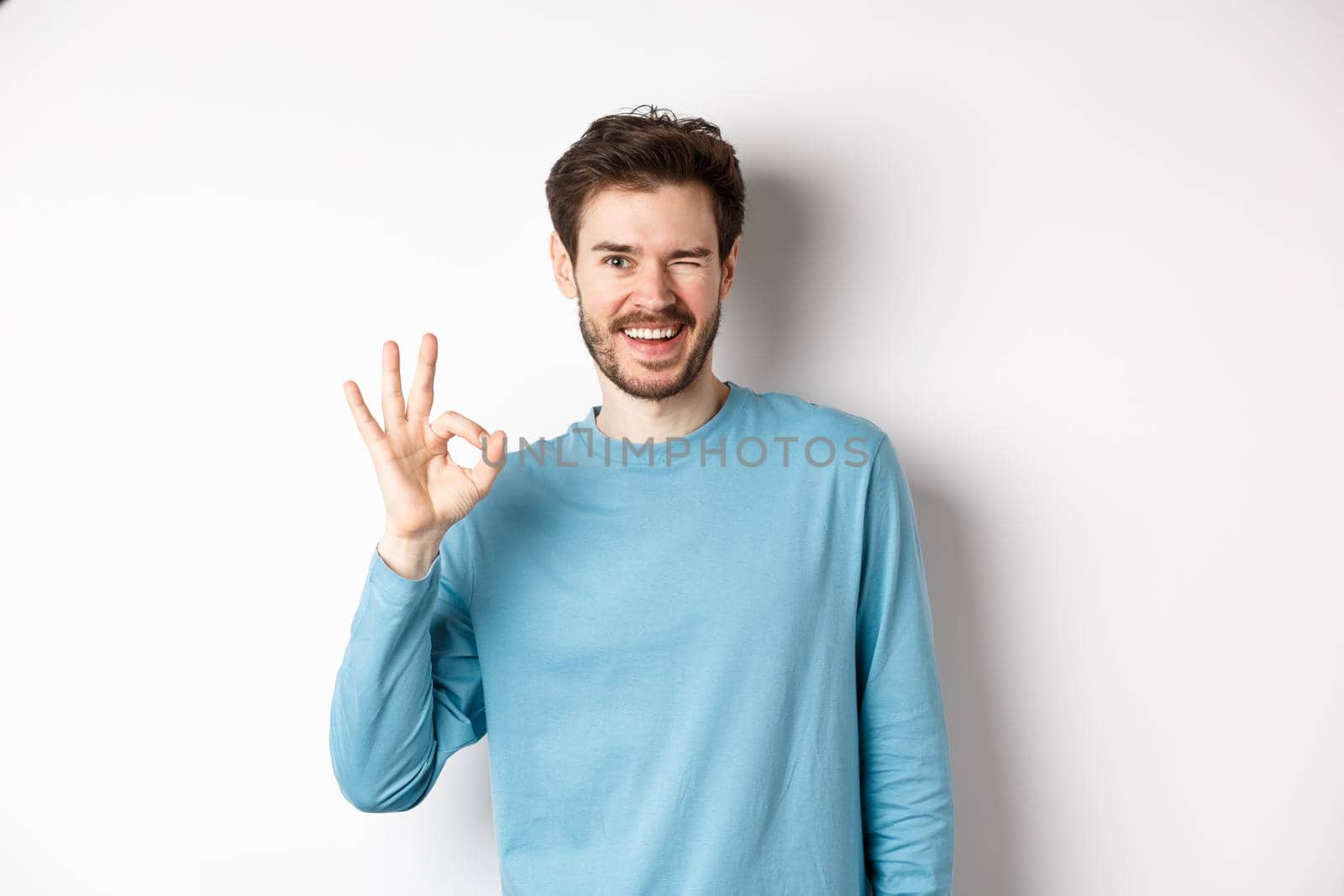 Handsome modern guy feeling confident, showing OK sign and winking at you, assure everything okay, standing over white background.