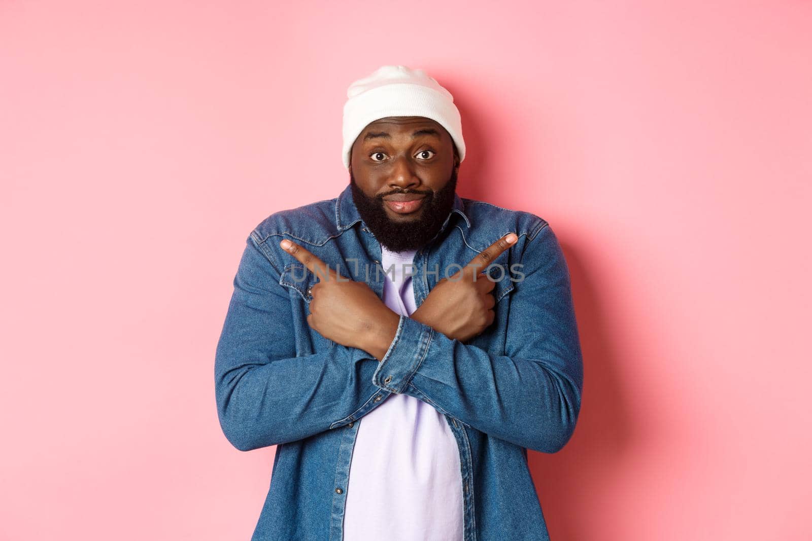 Confused Black man asking help with choice, pointing sideways and staring at camera, making decision, standing over pink background.