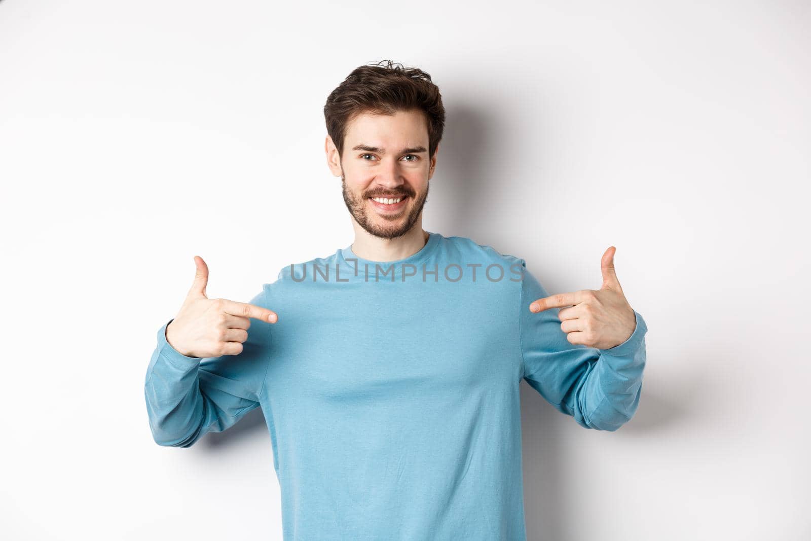 Handsome bearded man pointing fingers at center, self-promoting and looking confident, standing over white background, smiling at camera.