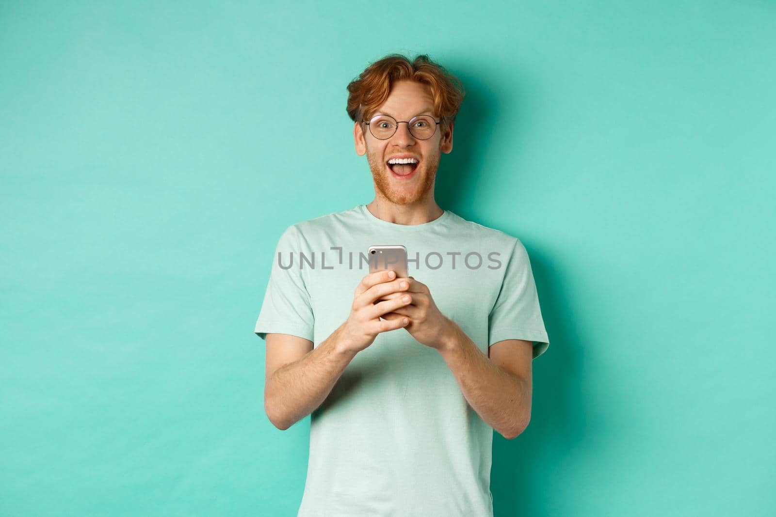 Surprised redhead man in glasses looking amazed at camera after reading promo offer on smartphone, standing against turquoise background.