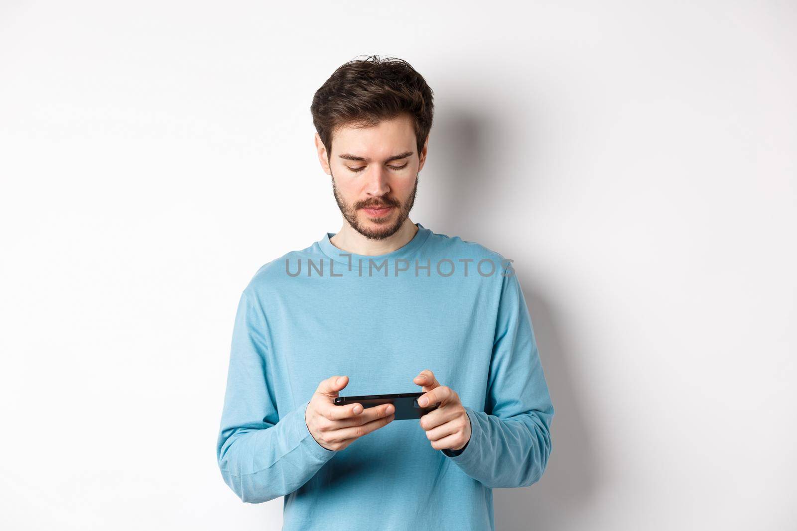 Male model playing video game on smartphone, looking serious at mobile screen, standing in sweatshirt over white background by Benzoix