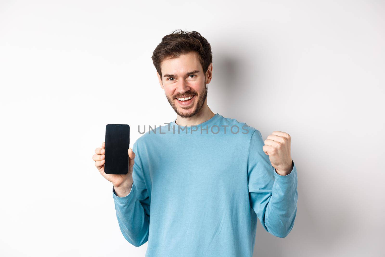 Image of happy man showing empty smartphone screen and celebrating, smiling with rejoice and fist pump, achieve online goal, standing over white background by Benzoix