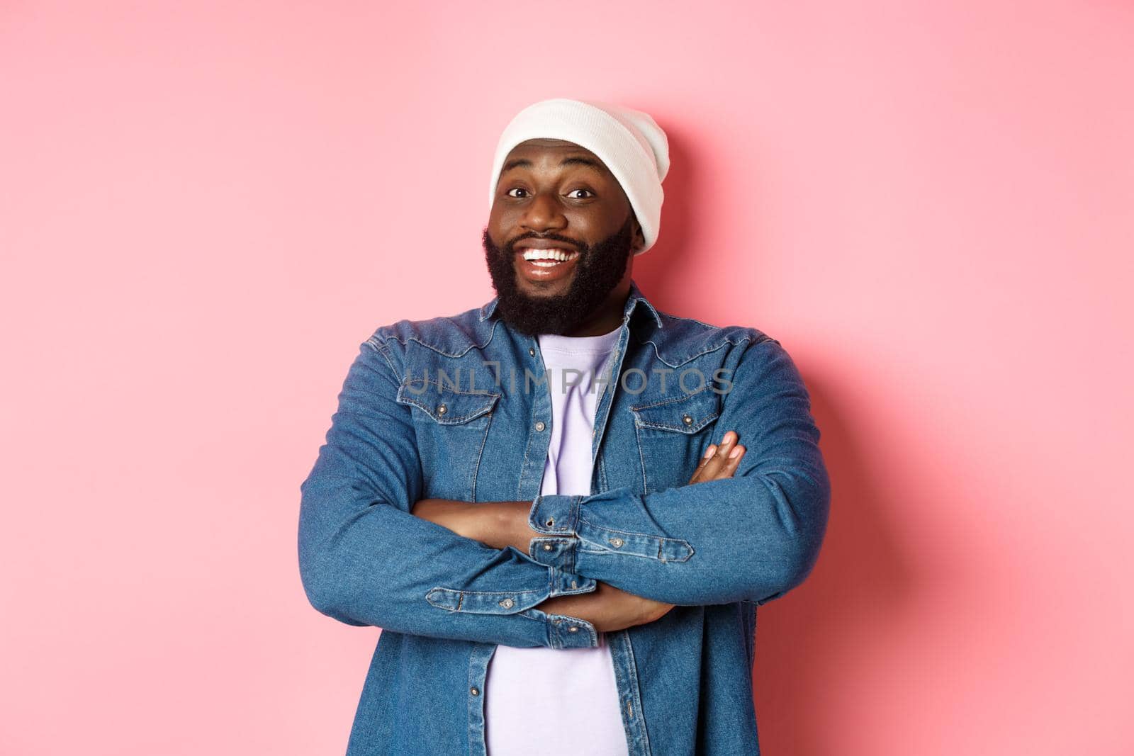 Happy Black man with beard, wearing beanie and denim shirt, looking intrigued and amused at camera, smiling with arms crossed on chest, pink background.