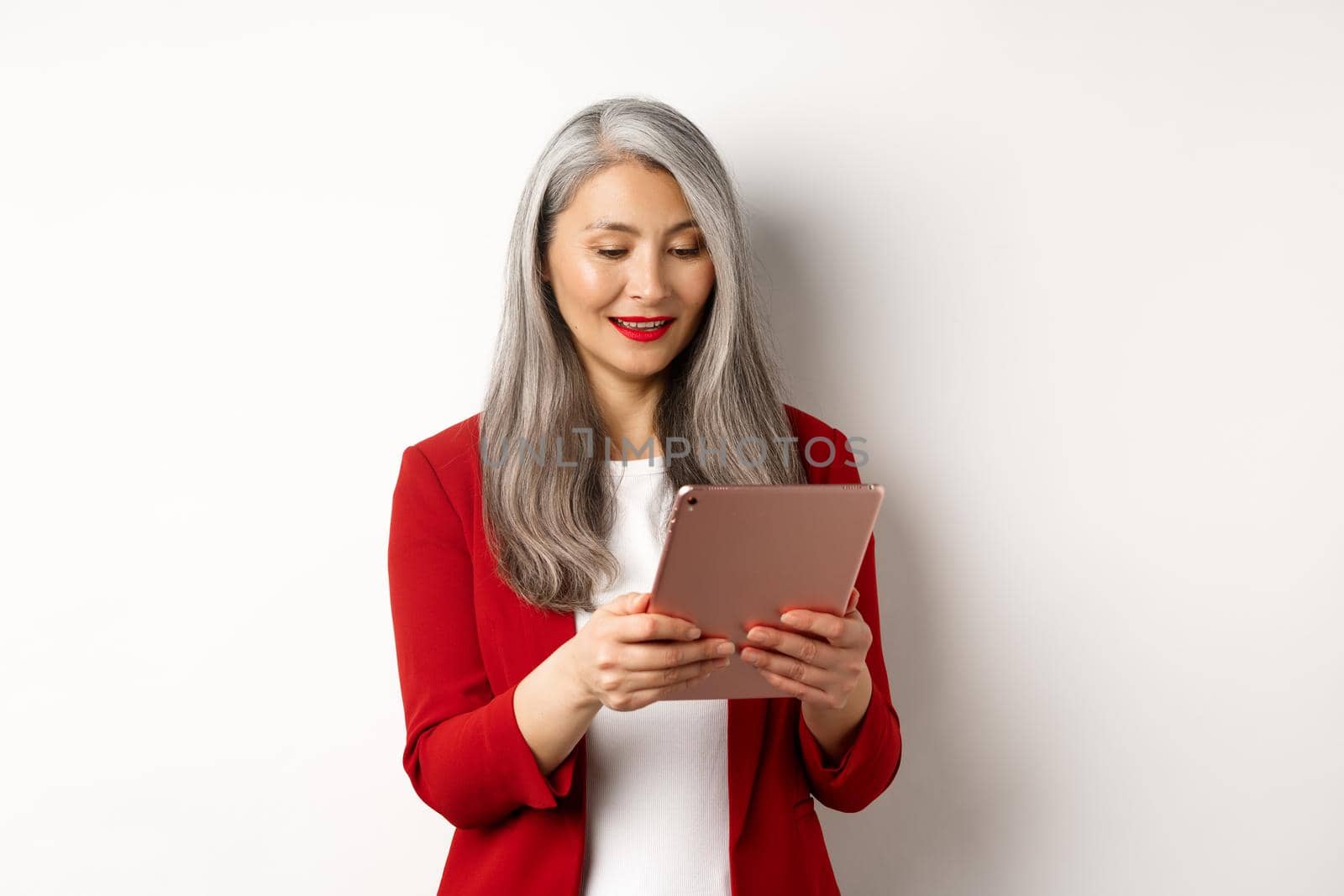 Business. Successful senior businesswoman working with digital tablet, reading screen and smiling, standing in elegant red blazer over white background by Benzoix