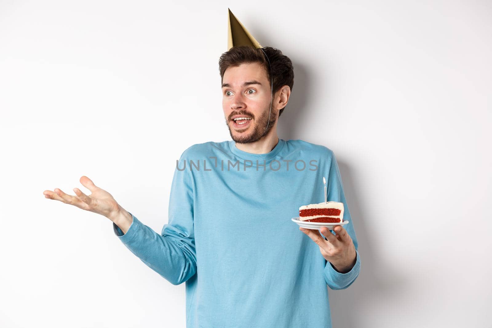 Celebration and holidays concept. Surprised man shrugging in disbelief, looking away, holding birthday cake and wearing party cone hat, white background by Benzoix