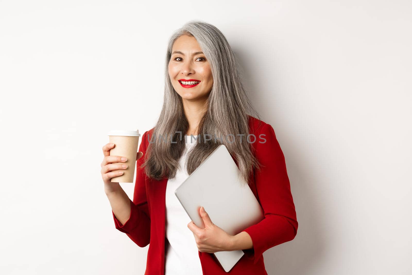 Business. Successful asian businesswoman with grey hair, wearing red blazer, drinking coffee and standing with laptop in hand, white background by Benzoix