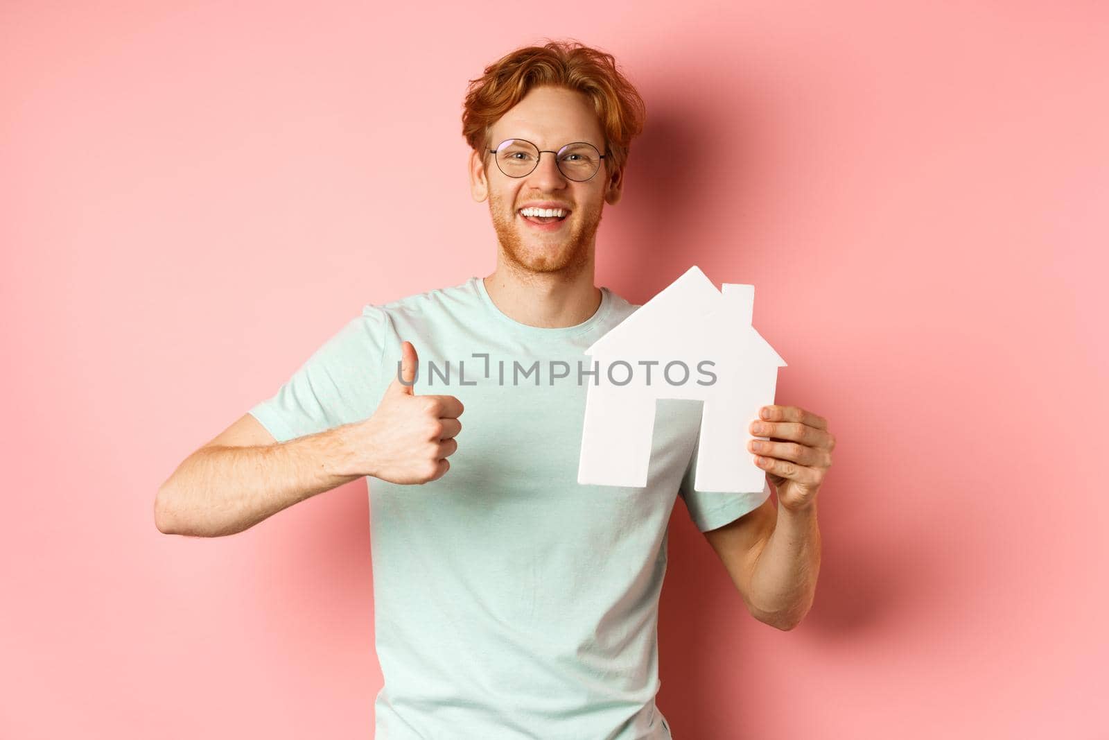 Real estate. Cheerful man in glasses and t-shirt recommending broker agency, showing paper house cutout and thumbs-up, standing over pink background by Benzoix